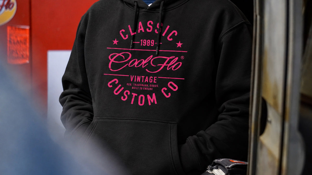Close-up of a model in a garage environment wearing a black  Cool Flo hoody with a bright pink graphic print on the front that reads 'CLASSIC COOL FLO VINTAGE CUSTOM CO 1989'.