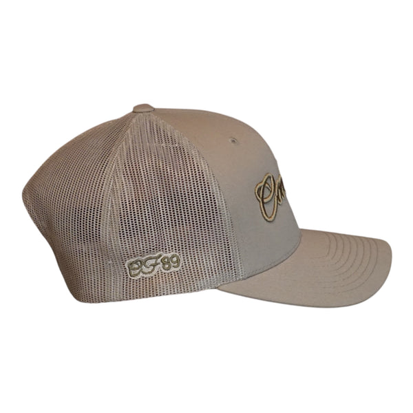 A khaki trucker cap with a mesh back, featuring a tonal embroidered logo on the side and a curved bronze logo on the front panel, shown against a plain white background.