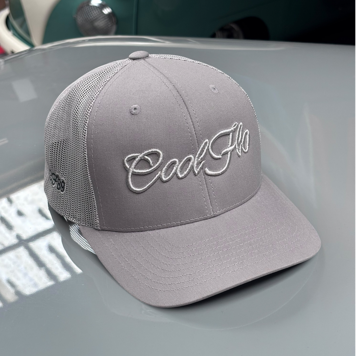 Front view of a Cool Flo silver-grey trucker cap featuring a raised 3D embroidered Script logo in tonal silver-grey and a curved visor.  Positioned on the bonnet of a grey classic car.