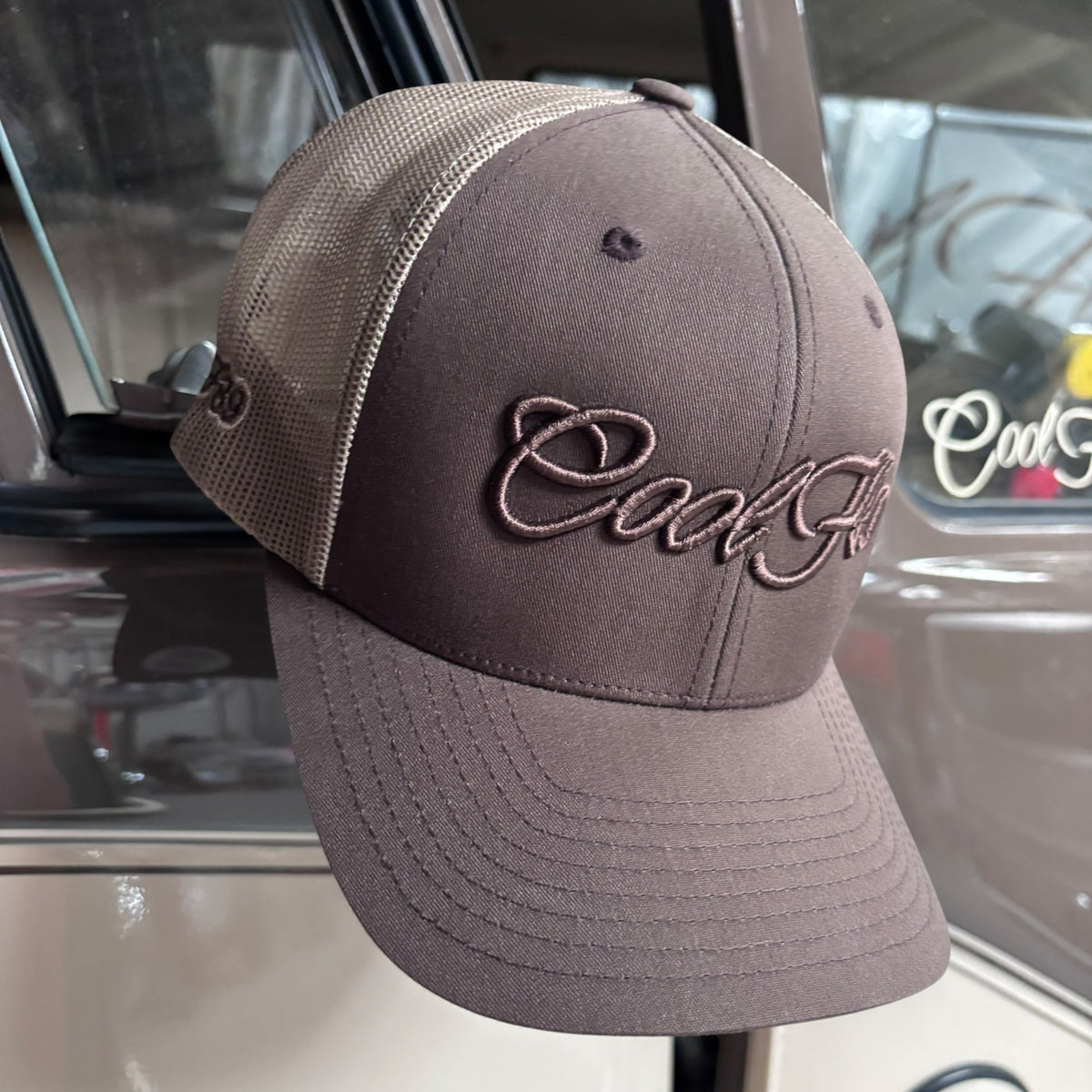 A brown and khaki trucker cap featuring the 'Cool Flo' script logo embroidered in brown across the front, displayed against a Cool Flo classic vehicle.