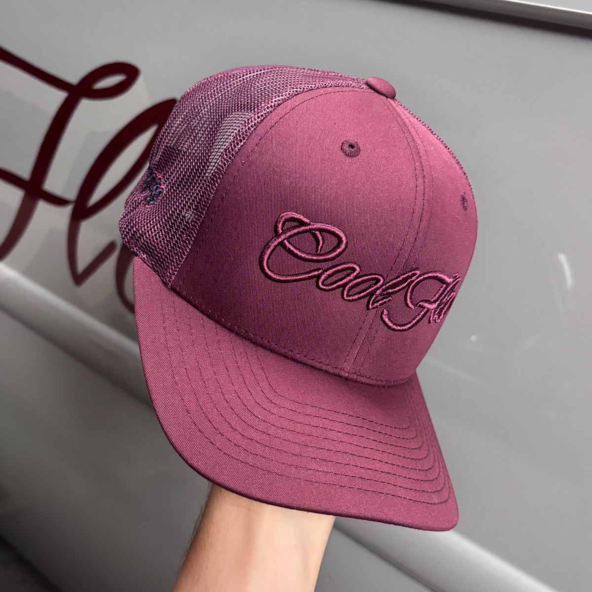 A burgundy trucker cap with 'Cool Flo' embroidered in matching burgundy script across the front, set against against a Cool Flo classic car.