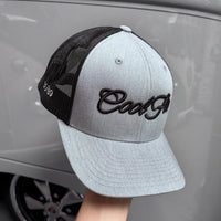 A heather grey trucker cap with black mesh side/back panels and black embroidered 'Cool Flo' script on the front, shown against a classic car.