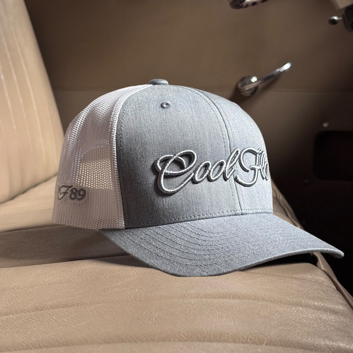 A heather grey trucker cap with white mesh side panels, and 'Cool Flo' embroidered in grey script across the front.