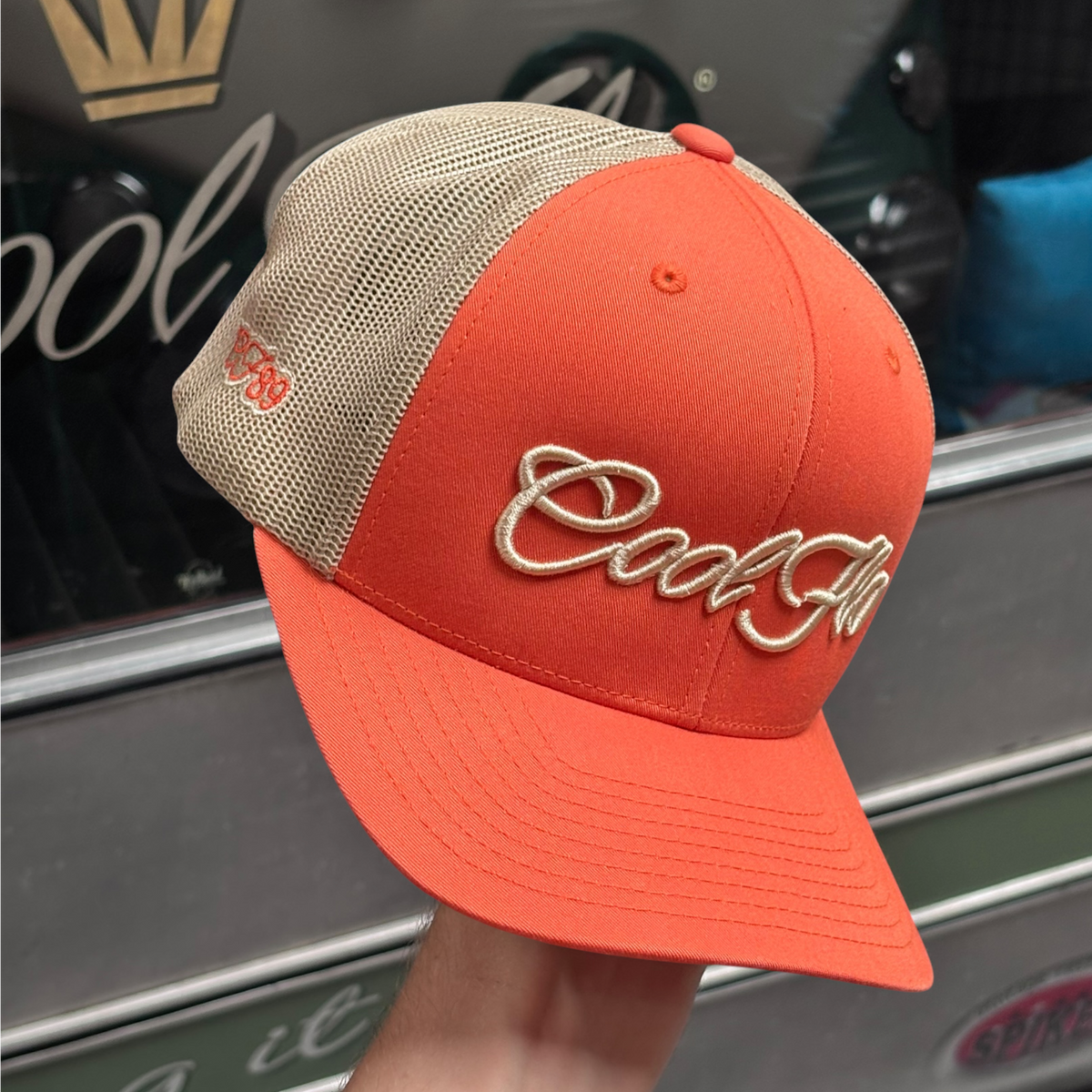 An orange trucker hat with khaki mesh back panels and 'Cool Flo' embroidered in yellow/gold script on the front, shown against a Cool Flo classic vehicle.