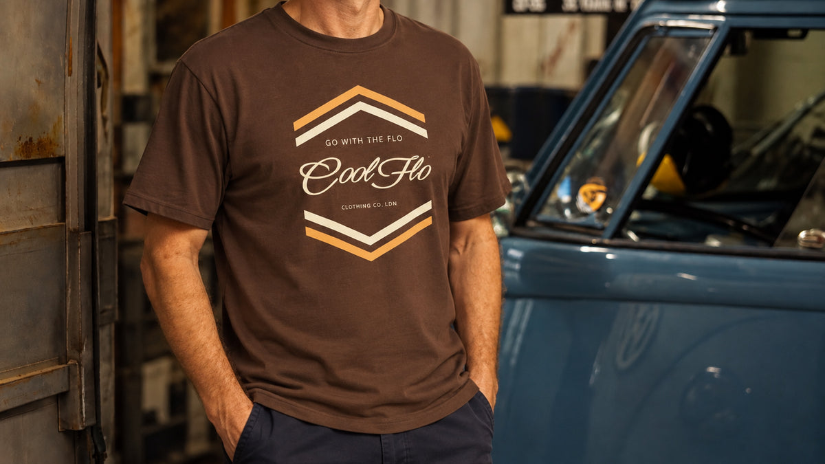 A model wearing a brown 'Chevron' graphic t-shirt with a 'Cool Flo' logo, standing in a garage next to a blue vintage VW panel van.