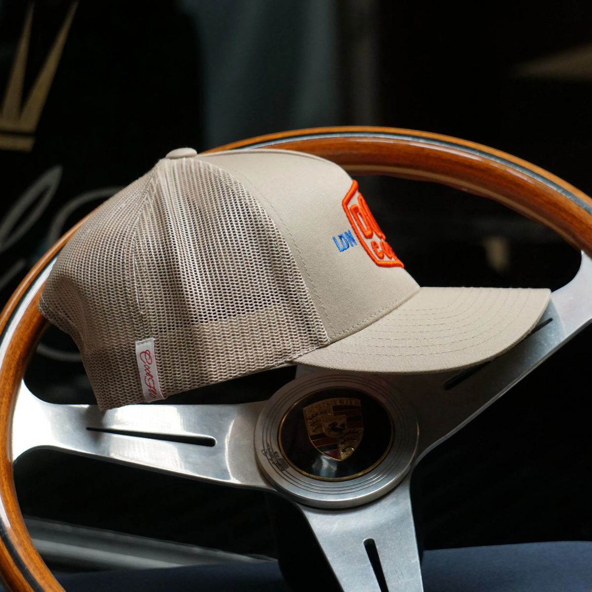 Side-on view of Douze Coupes khaki trucker cap  with red and blue embroidery, sitting on a classic wood and metal steering wheel.
