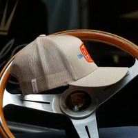 Side-on view of Douze Coupes khaki trucker cap  with red and blue embroidery, sitting on a classic wood and metal steering wheel.