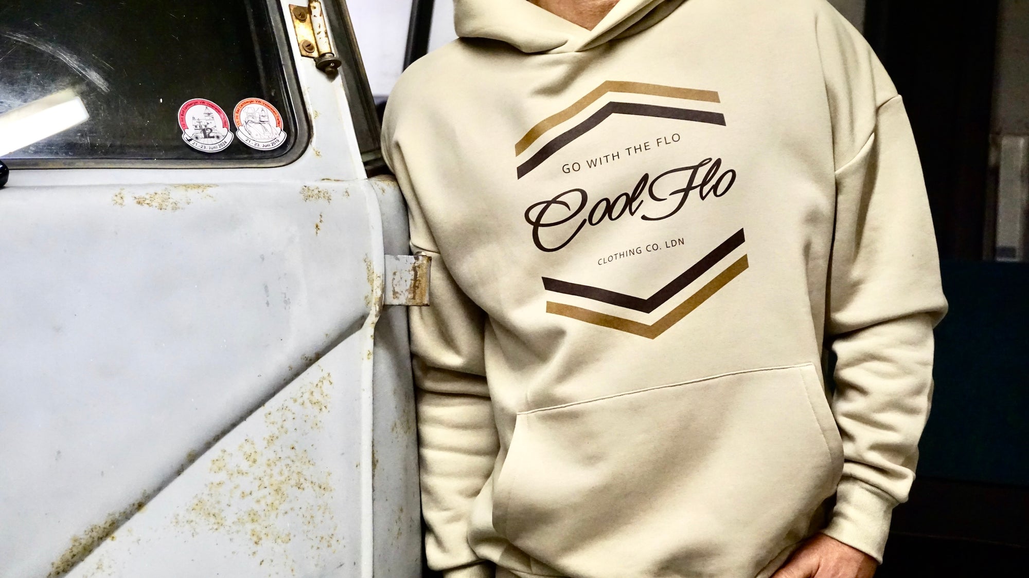 Cropped image of model wearing a stone colour hoody with light and dark brown chevron Cool Flo Go With the Flo printed design, stood by a vintage grey VW bus.