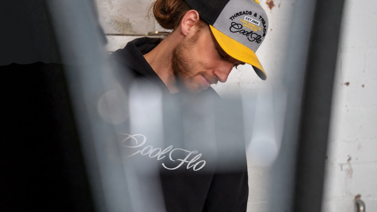 Male model in garage environment, looking down wearing a yellow, grey and black embroidered Threads and Treads Cool Flo cap and black Cool Flo script logo hoody. 