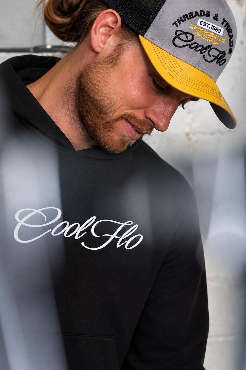 Male model in garage environment, looking down wearing a yellow, grey and black embroidered Threads and Treads Cool Flo cap and black Cool Flo script logo hoody. 