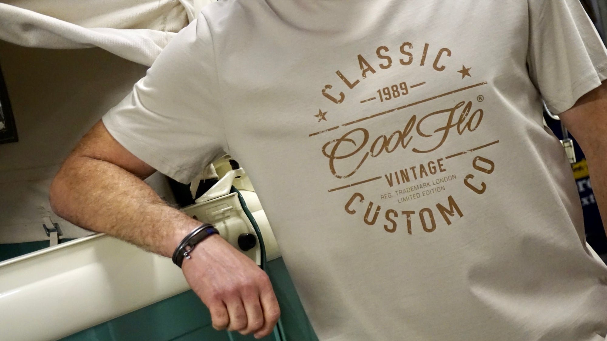 Close-up of model leaning on vintage cream and green VW pick-up truck, wearing a stone colour Cool Flo t-shirt with 'Classic Cool Flo Vintage Custom Co' text design printed in brown on the chest.