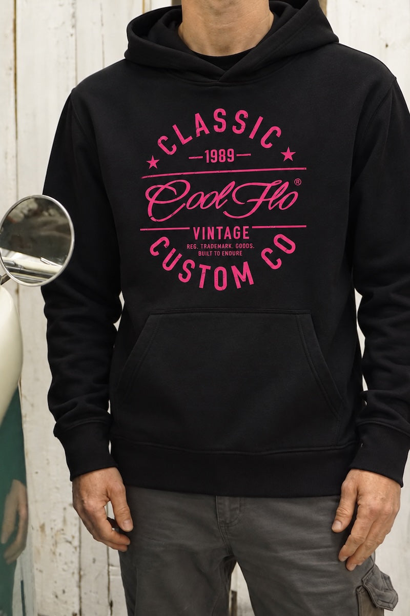 Close-up of a model stood next to a green and cream vintage VW pick-up truck wearing a black  Cool Flo hoody with a bright pink graphic print on the front that reads 'CLASSIC COOL FLO VINTAGE CUSTOM CO 1989'.