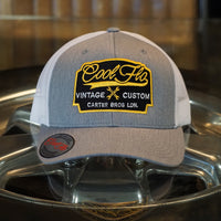 Cool Flo Vintage Custom. Grey and white trucker cap with embroidered yellow, black and white badge with cross-spanners. Carter Bros. London.