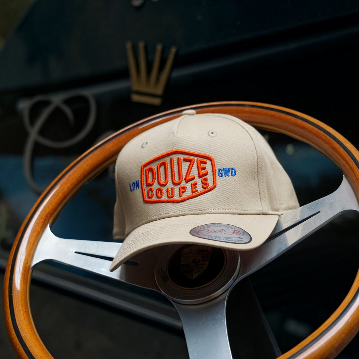 Douze Coupes khaki baseball cap  with red and blue embroidery, sitting on a classic wood and metal steering wheel.