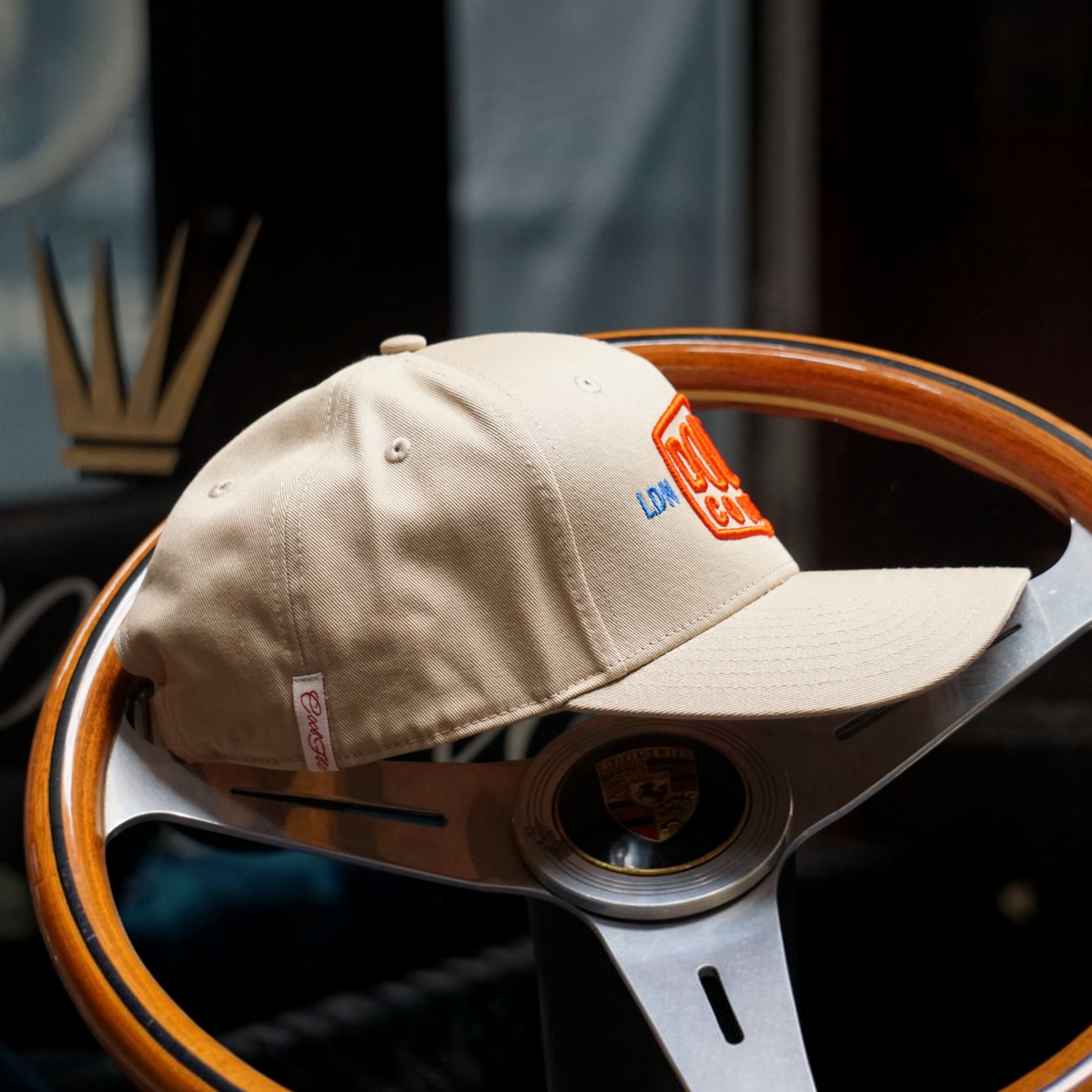 Side-on view of Douze Coupes khaki baseball cap  with red and blue embroidery, sitting on a classic wood and metal steering wheel.