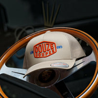 Douze Coupes khaki baseball cap  with red and blue embroidery, sitting on a classic wood and metal steering wheel.